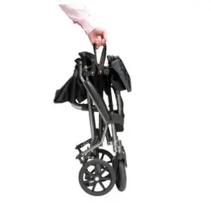 A person holds the Drive TraveLite Aluminium Transport Chair, folded by its handle, against a plain white background.