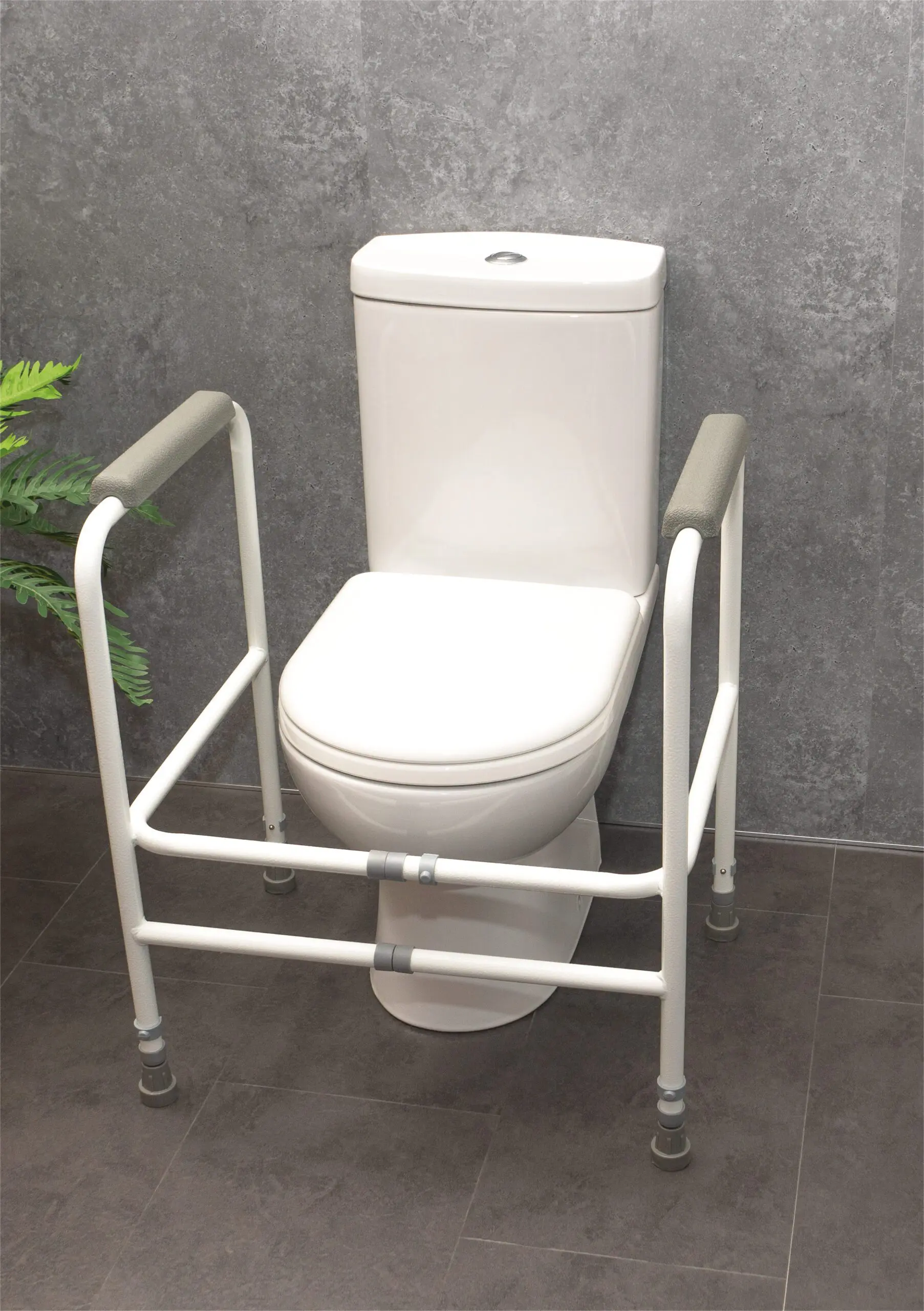 The Broadstairs Toilet Frame with Adjustable Height and Width, featuring metal arms, is placed around a white toilet set against a grey tiled wall on a dark floor.
