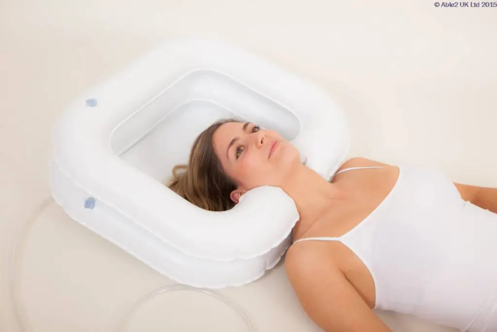 A woman lies on her back with her head resting in the ATLANTIS DELUXE INFLATABLE SHAMPOO RING, a white basin designed for washing hair in bed.