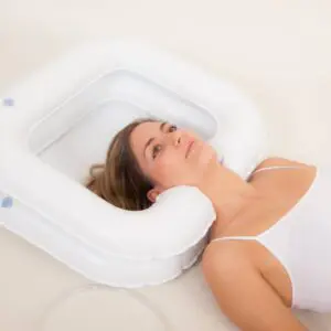 A woman lies on her back with her head resting in the ATLANTIS DELUXE INFLATABLE SHAMPOO RING, a white basin designed for washing hair in bed.