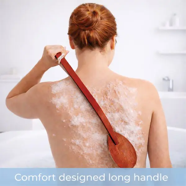 A person with red hair uses the Long-Handled Wooden Bath Brush to scrub their back whilst sitting in a bath filled with lathered soap.