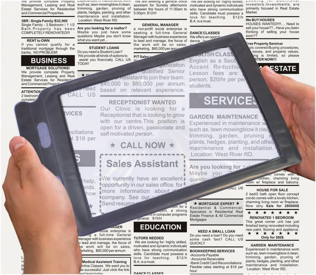 Hands use the Sheet Magnifier over a newspaper’s classified adverts, enlarging job listings such as receptionist and sales assistant posts.