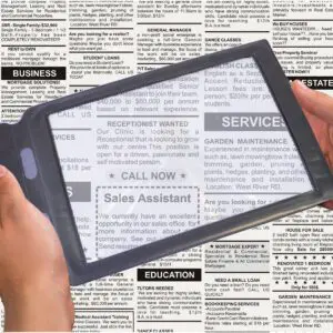 Hands use the Sheet Magnifier over a newspaper’s classified adverts, enlarging job listings such as receptionist and sales assistant posts.
