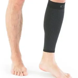 A person stands barefoot, wearing a black compression sleeve on their right calf, whilst their left leg is bare against a white background.