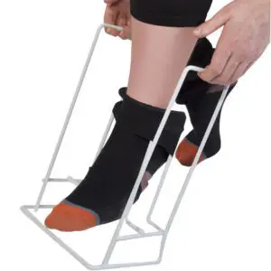 A person uses the Rigid Sock & Stocking Helper to put on socks by holding the frame with both hands and sliding their foot into the sock.