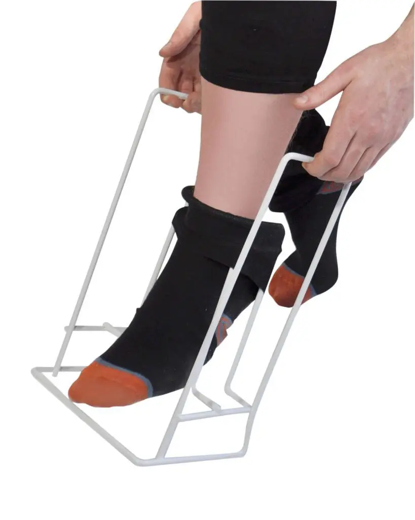 A person uses the Rigid Sock & Stocking Helper to put on socks by holding the frame with both hands and sliding their foot into the sock.