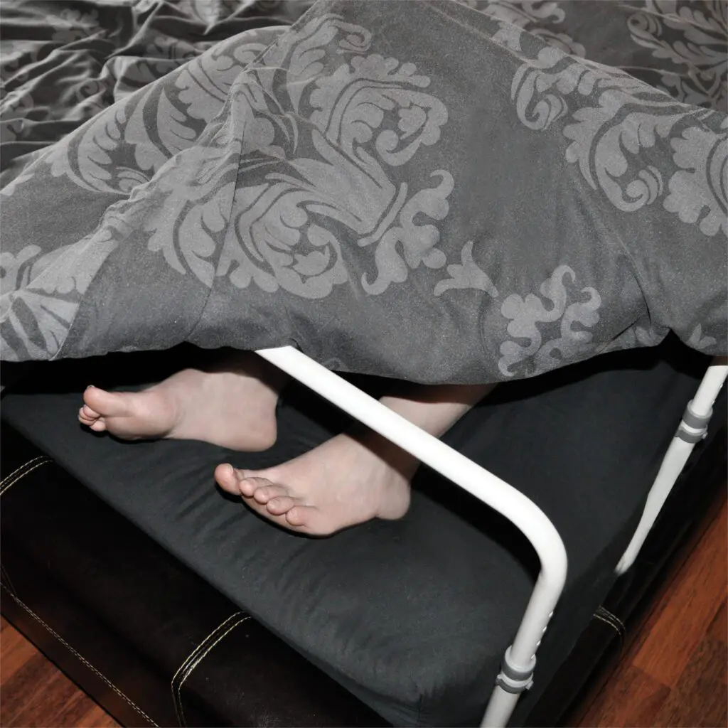 Two bare feet peek out from under a grey patterned blanket on a fold-out bed with a white metal frame, featuring the Height Adjustable Bed Cradle for extra comfort.