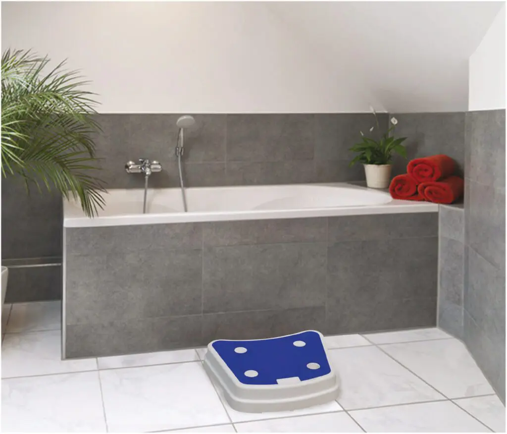 A grey-tiled bathroom with a bathtub, handheld showerhead, green plant, red towels, and a Plastic Stack-able Bath Step in blue and white on the floor.