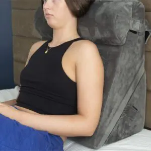 A person sits upright on a bed, leaning against the Luxury Folding Adjustable Bed Wedge Pillow, which is large, angled, grey, and features a cylindrical neck support.