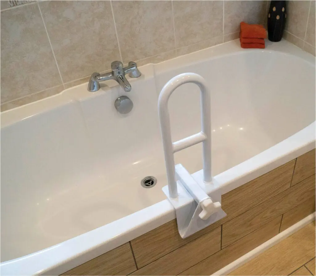 A white bath features a Bath Grab Rail mounted on the edge, a tap on the tiled wall, and two orange towels stacked in the corner.