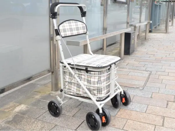 A checked-patterned wheeled shopping trolley with a seat is parked on a pavement near a glass bus shelter.