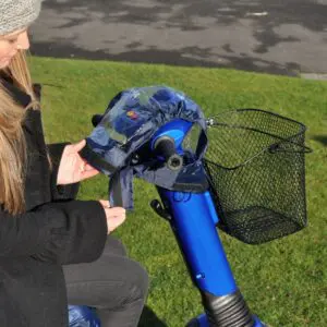 A woman on a mobility scooter attaches the blue Splash Scooter Control Panel Cover to the scooter's control panel and handlebars.