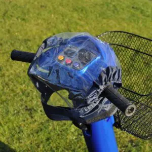 Electric scooter handlebars with a transparent plastic cover over the control panel and a metal basket attached at the front, positioned on grass.