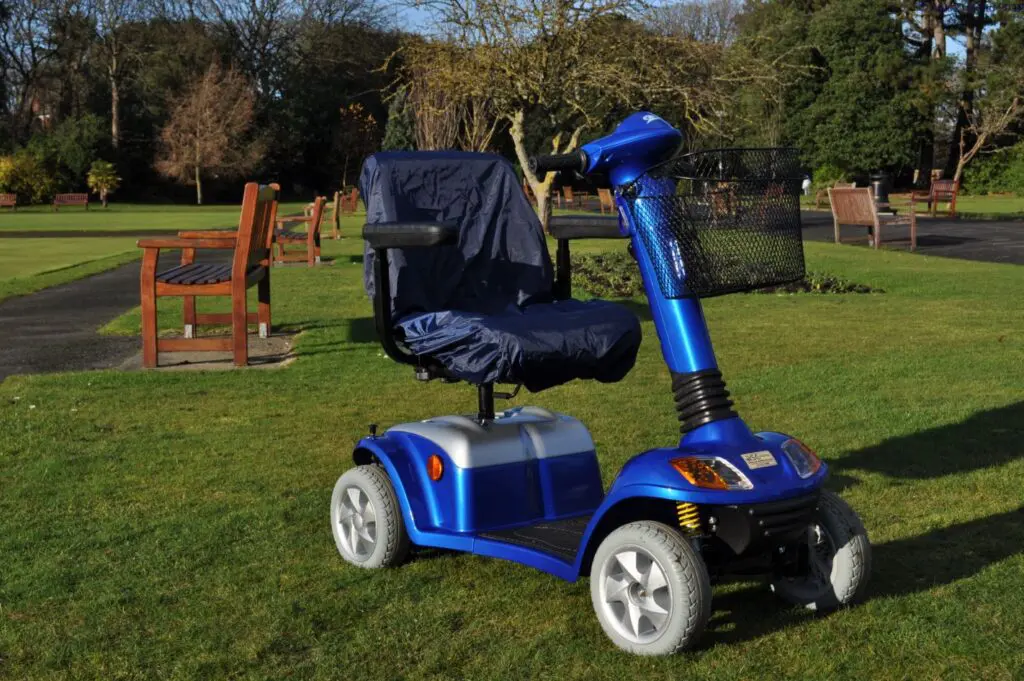 A blue mobility scooter with a covered seat is parked on grass in a park with benches and trees in the background.