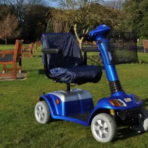 A blue mobility scooter with a covered seat is parked on grass in a park with benches and trees in the background.