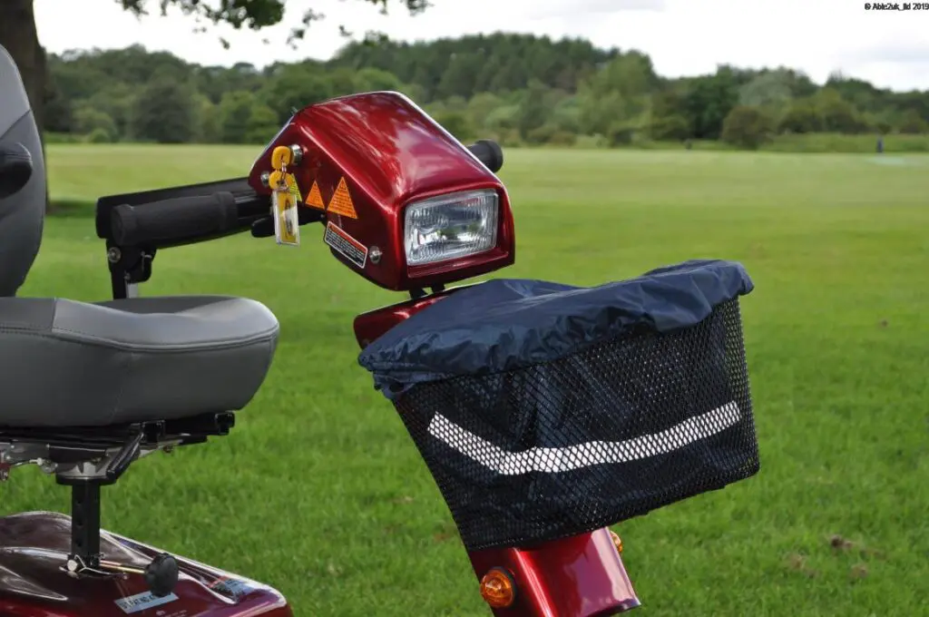 Close-up of a red mobility scooter parked on grass, featuring the Splash Scooter Basket Liner Bag & Cover in the mesh basket, along with the handlebar, headlamp, seat, and greenery in the background.