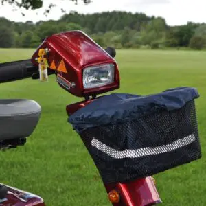 Close-up of a red mobility scooter parked on grass, featuring the Splash Scooter Basket Liner Bag & Cover in the mesh basket, along with the handlebar, headlamp, seat, and greenery in the background.