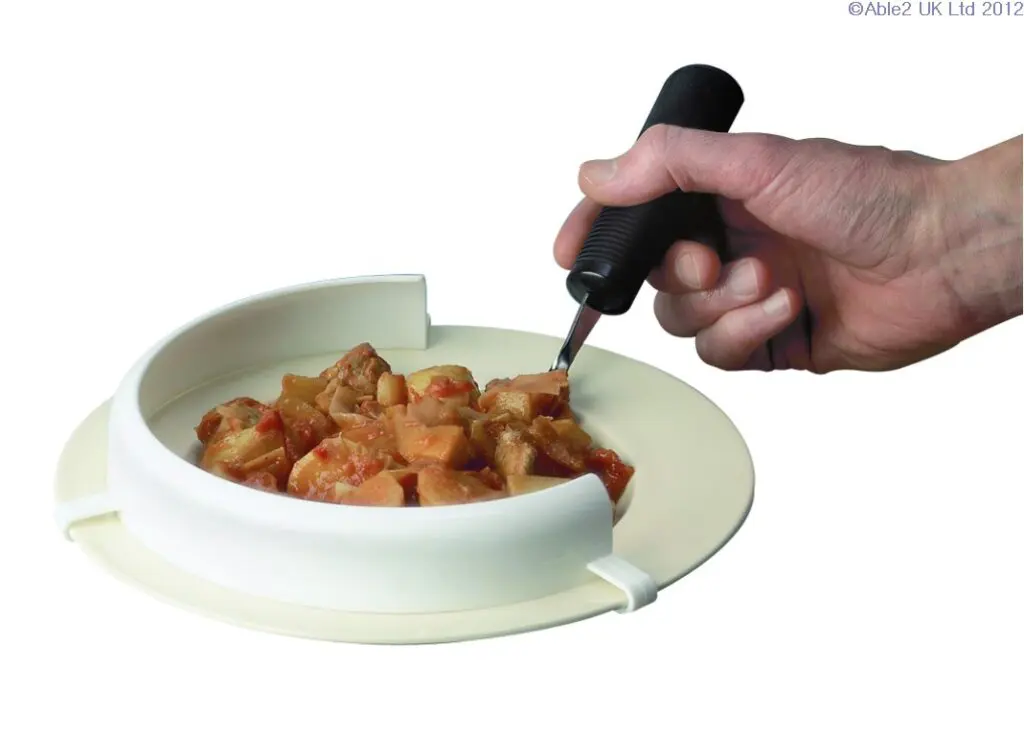 A hand uses a fork to eat food from a plate fitted with the Food Guard, which features a curved, raised edge designed to assist with scooping.