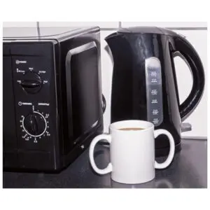 A black electric kettle with steam rising sits on a kitchen worktop next to a microwave, with two white mugs in front, one containing a hot drink.