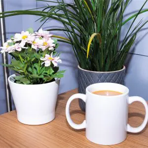 A white mug of coffee sits on a wooden table next to a potted plant with pink flowers and another green potted plant.