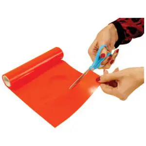 A person uses blue-handled scissors to cut a sheet of glossy red plastic film from a roll.