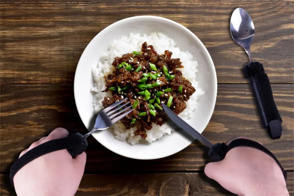 A person with prosthetic hands uses the Bendable Cutlery Set (6 piece) to hold a fork and knife, getting ready to enjoy a meal of white rice with saucy meat and chopped spring onions.