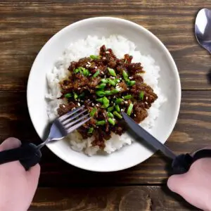 A person with prosthetic hands uses the Bendable Cutlery Set (6 piece) to hold a fork and knife, getting ready to enjoy a meal of white rice with saucy meat and chopped spring onions.