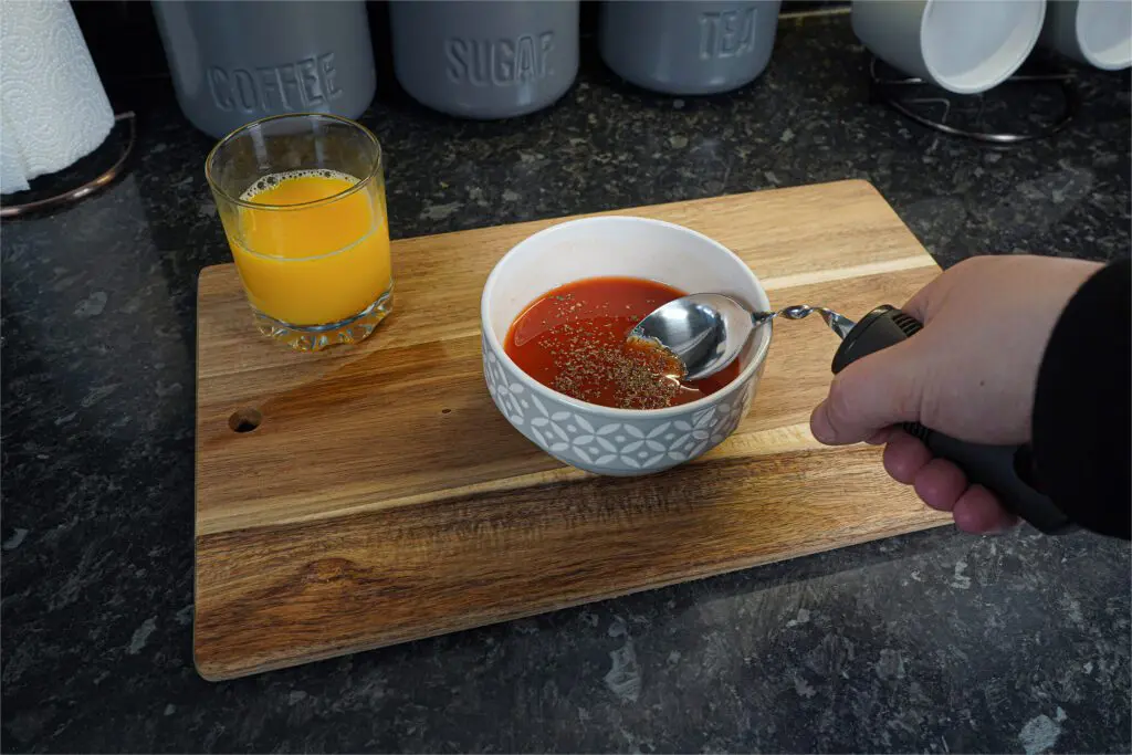 A hand holds a Large Handled Bendable Spoon over a bowl of tomato soup with herbs on a wooden board, next to a glass of orange juice.