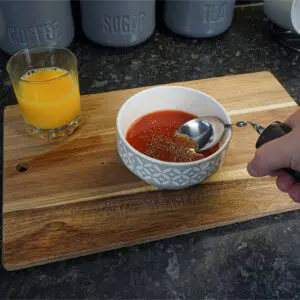 A hand holds a Large Handled Bendable Spoon over a bowl of tomato soup with herbs on a wooden board, next to a glass of orange juice.