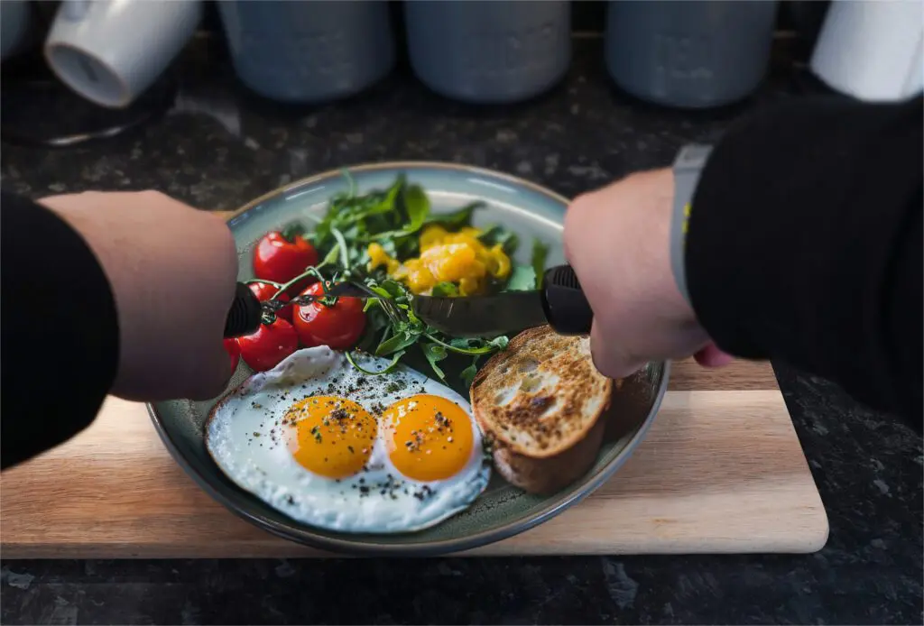 A person uses the Large Handled Bendable Cutlery Set to cut food on a plate with two fried eggs, cherry tomatoes, greens, sliced yellow pepper, and toasted bread on a wooden board.
