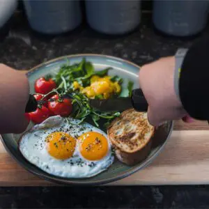 A person uses the Large Handled Bendable Cutlery Set to cut food on a plate with two fried eggs, cherry tomatoes, greens, sliced yellow pepper, and toasted bread on a wooden board.
