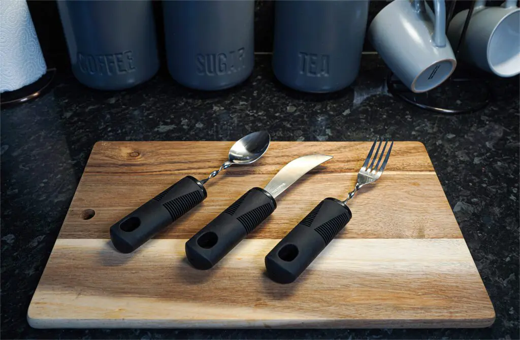 The Large Handled Bendable Cutlery Set, featuring thick black handles, is displayed on a wooden chopping board with kitchen canisters and mugs in the background.