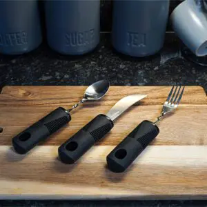 The Large Handled Bendable Cutlery Set, featuring thick black handles, is displayed on a wooden chopping board with kitchen canisters and mugs in the background.
