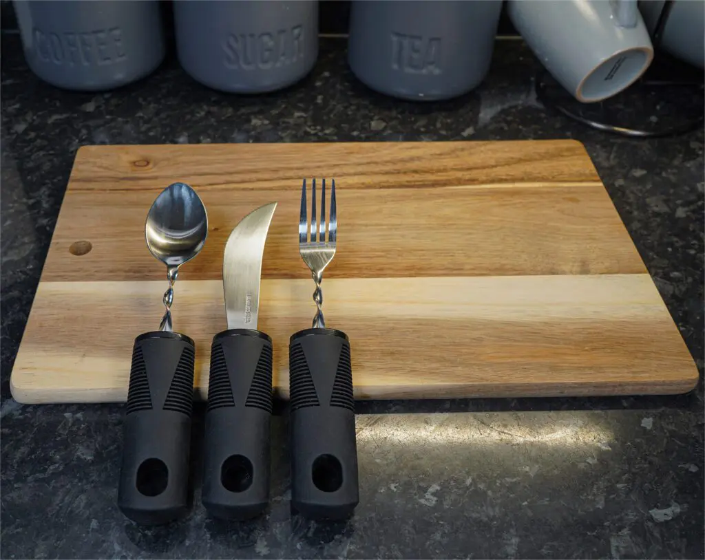 The Large Handled Bendable Cutlery Set, featuring a spoon, knife, and fork with black handles, is arranged on a wooden chopping board with coffee, sugar, and tea containers in the background.