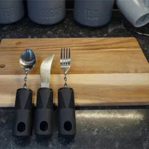The Large Handled Bendable Cutlery Set, featuring a spoon, knife, and fork with black handles, is arranged on a wooden chopping board with coffee, sugar, and tea containers in the background.