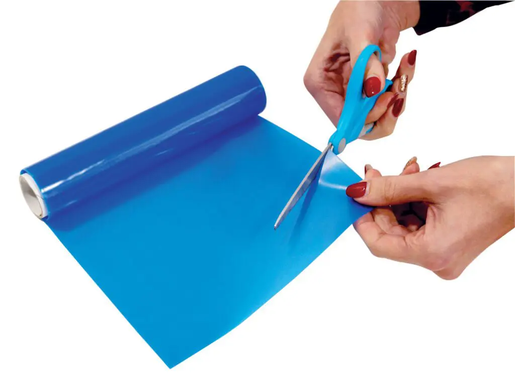 Person cutting a sheet of blue plastic film from a roll using blue-handled scissors against a white background.