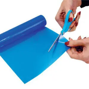 Person cutting a sheet of blue plastic film from a roll using blue-handled scissors against a white background.