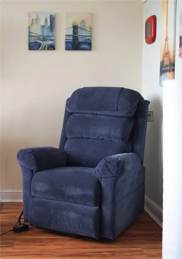 A blue upholstered reclining chair sits in the corner of a room with light walls, wooden flooring, and framed artwork on the wall above.