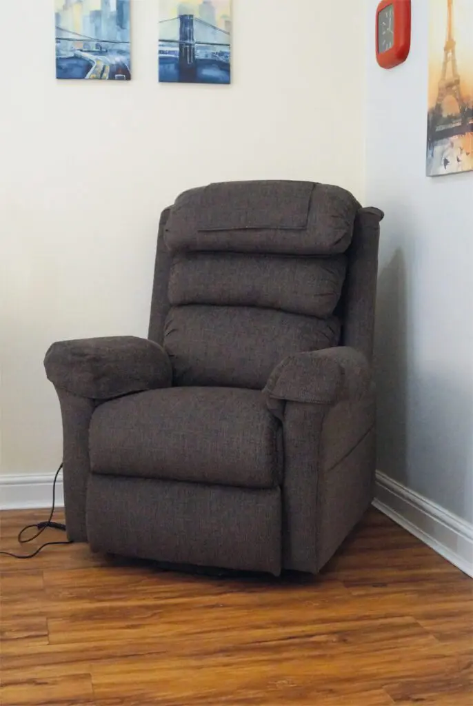 A dark grey recliner chair is positioned in the corner of a room with wooden flooring. Two paintings and a red wall clock are hung on the walls above and beside the chair.
