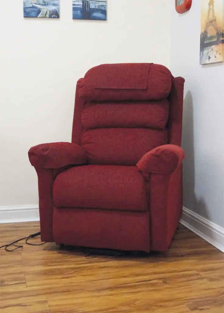 A red upholstered reclining chair sits on a wooden floor in the corner of a room, with paintings and power leads visible nearby.