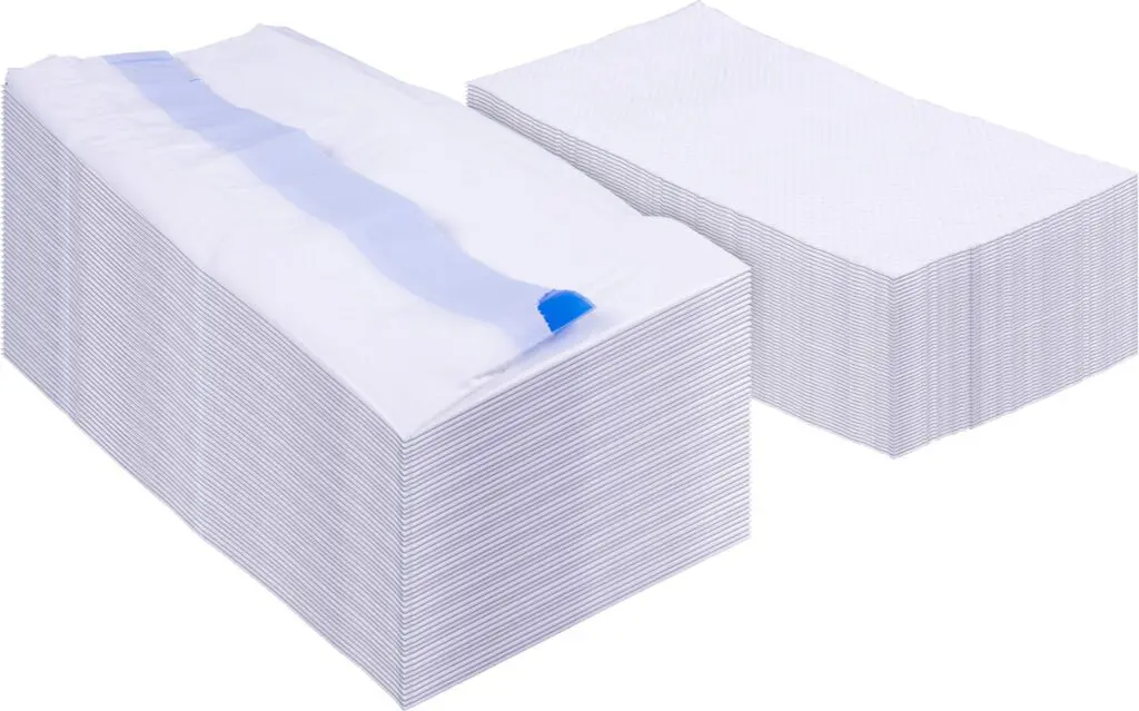 Two large stacks of rectangular white Commode Liners with Absorbent Pads (Box of 50), one stack partially covered by a single unfolded pad showing its blue adhesive strip.