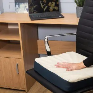 A hand presses down on the Gel Comfort Cushion, a white seat pad placed on a black office chair in front of a wooden desk with a laptop, plant, and framed Eiffel Tower picture.