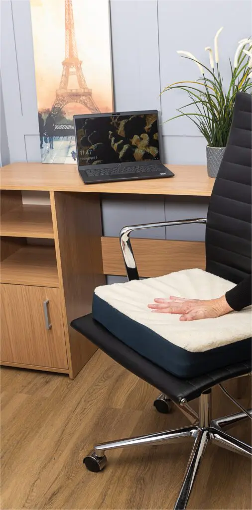 A hand presses down on the Gel Comfort Cushion, a white seat pad placed on a black office chair in front of a wooden desk with a laptop, plant, and framed Eiffel Tower picture.