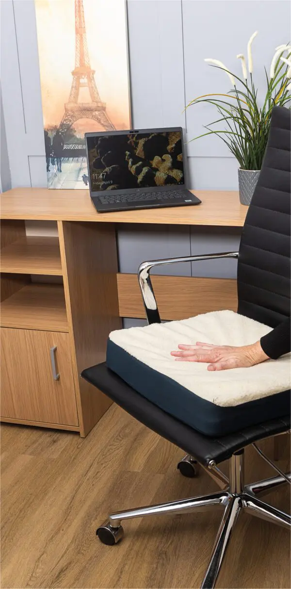 A hand presses down on the Gel Comfort Cushion, a white seat pad placed on a black office chair in front of a wooden desk with a laptop, plant, and framed Eiffel Tower picture.