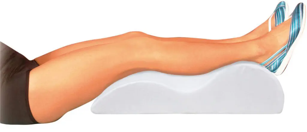 A person rests on their back with legs elevated on the Foam Leg Raiser, wearing black shorts and blue striped shoes.