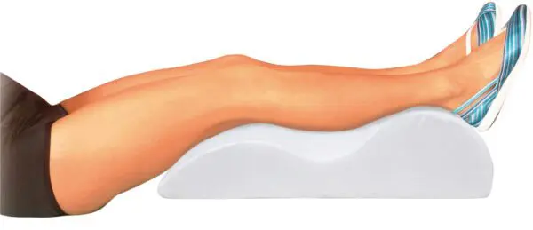 A person rests on their back with legs elevated on the Foam Leg Raiser, wearing black shorts and blue striped shoes.