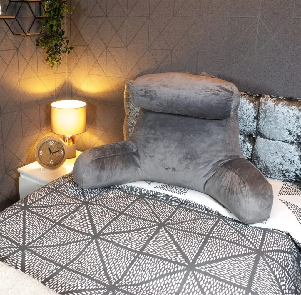 A reading pillow with head rest and back support in grey is placed on a patterned bedspread next to a bedside table holding a lamp, clock, and plant.
