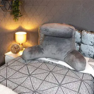 A reading pillow with head rest and back support in grey is placed on a patterned bedspread next to a bedside table holding a lamp, clock, and plant.