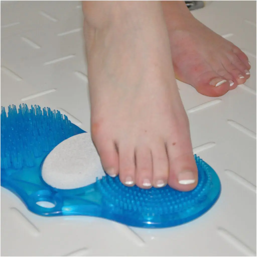 A person uses the Foot Cleaner with pumice, featuring blue bristles and a pumice stone, to clean their foot on a white textured shower floor.