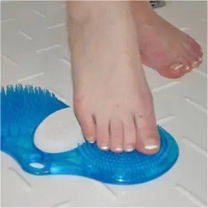 A person uses the Foot Cleaner with pumice, featuring blue bristles and a pumice stone, to clean their foot on a white textured shower floor.